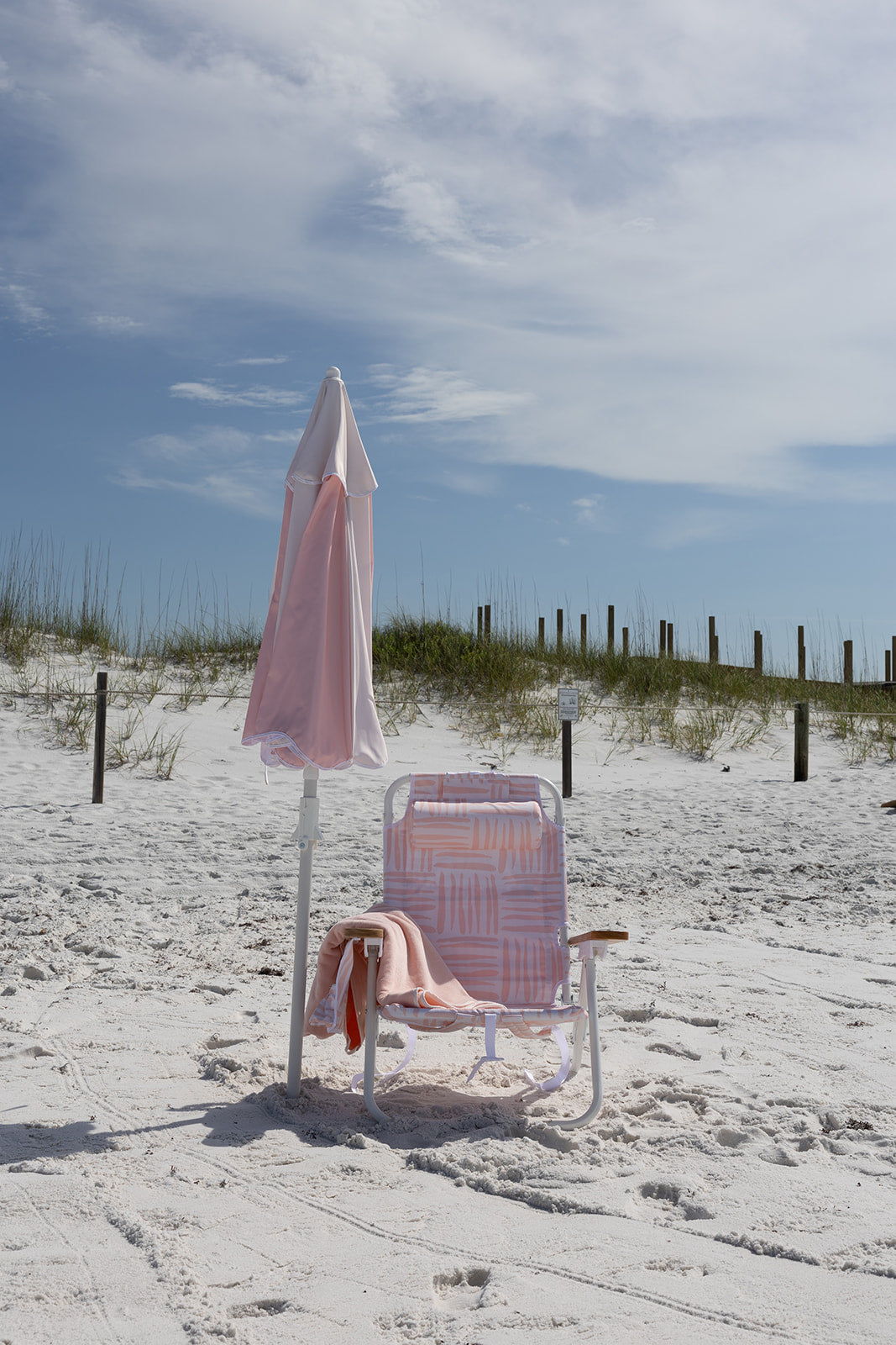 Pink patterned beach chair with pink towel and blush rose colored umbrella on a sandy beach with grassy dunes and blue sky.