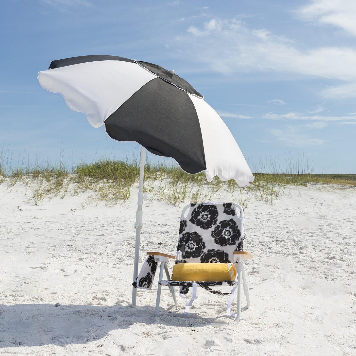 Beach chair with black and white flowers, with a yellow towel on it, a under a black and white umbrella on a sandy beach.