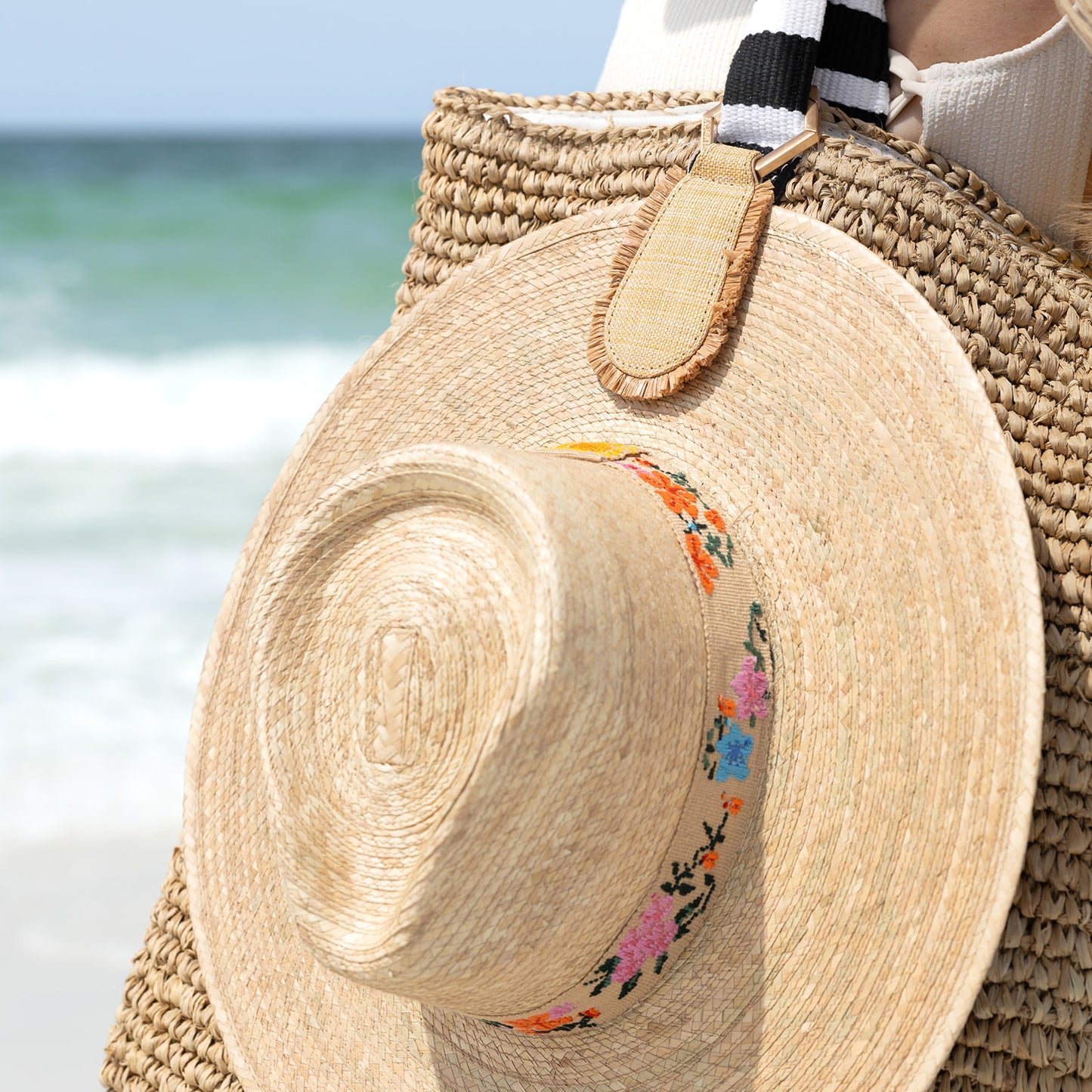 A natural straw hat with floral band held by a raffia hat clipped to a woven bag against a beach background