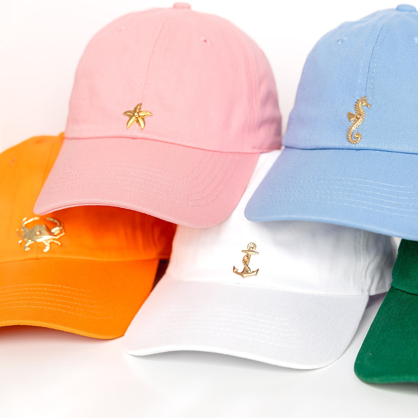 Five colorful baseball caps displayed on a white background: pink cap with gold starfish, powder blue cap with gold seahorse, white cap with gold anchor, orange cap with gold crab, and green cap.