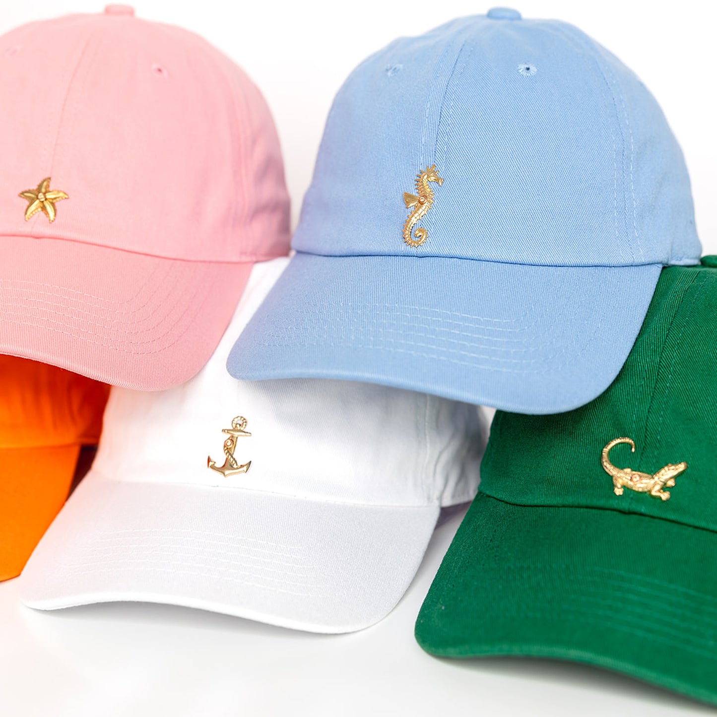 Five colorful baseball caps displayed on a white background: pink cap with gold starfish, powder blue cap with gold seahorse, white cap with gold anchor, orange cap with gold crab, and green cap.