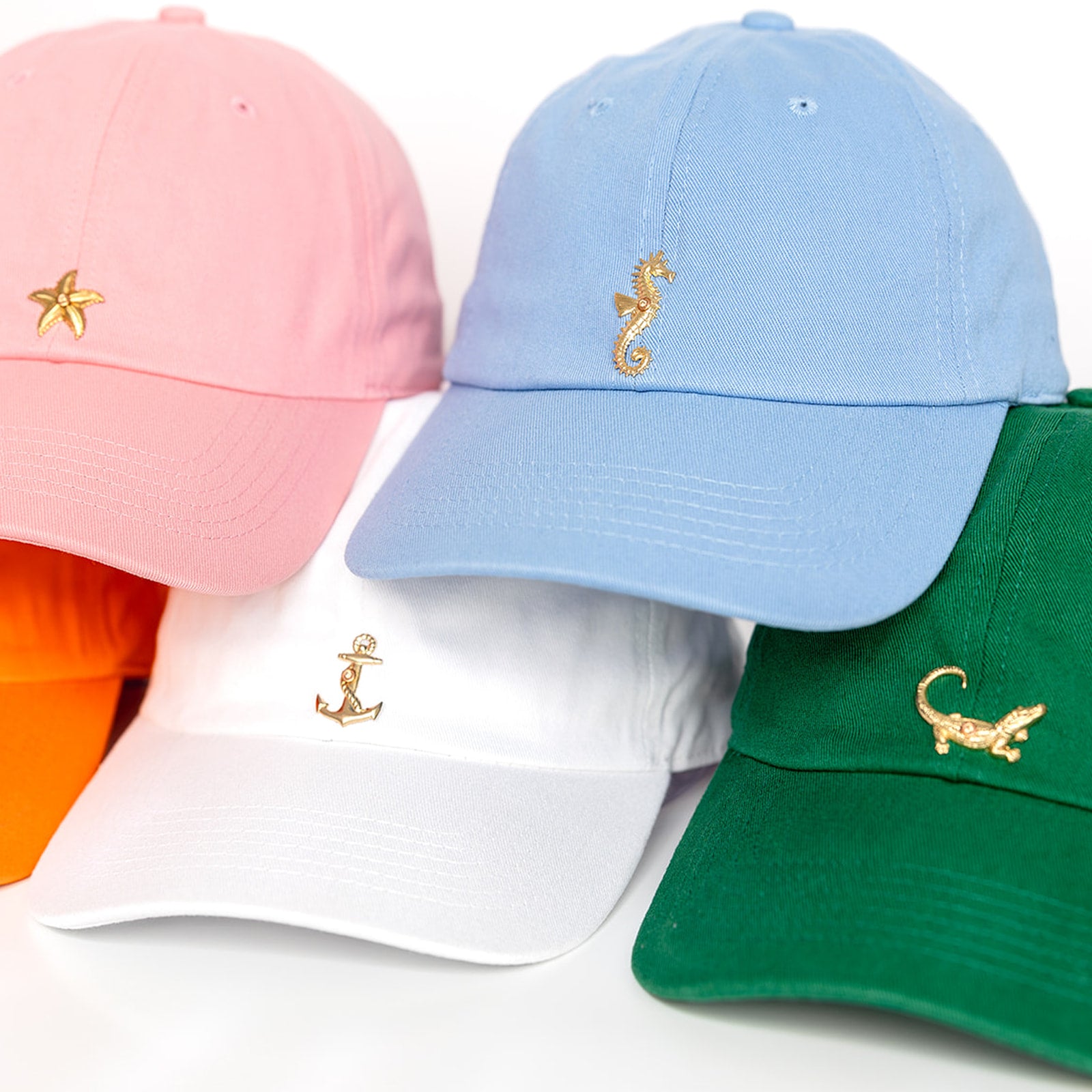 Five colorful baseball hats displayed on a white background: pink cap with gold starfish,  powder blue cap with gold seahorse, white hat with gold anchor, orange cap with gold crab, and green cap.