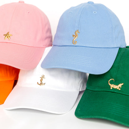 Five colorful baseball caps displayed on a white background: pink cap with gold starfish,  powder blue cap with gold seahorse, white cap with gold anchor, orange cap with gold crab, and green cap.