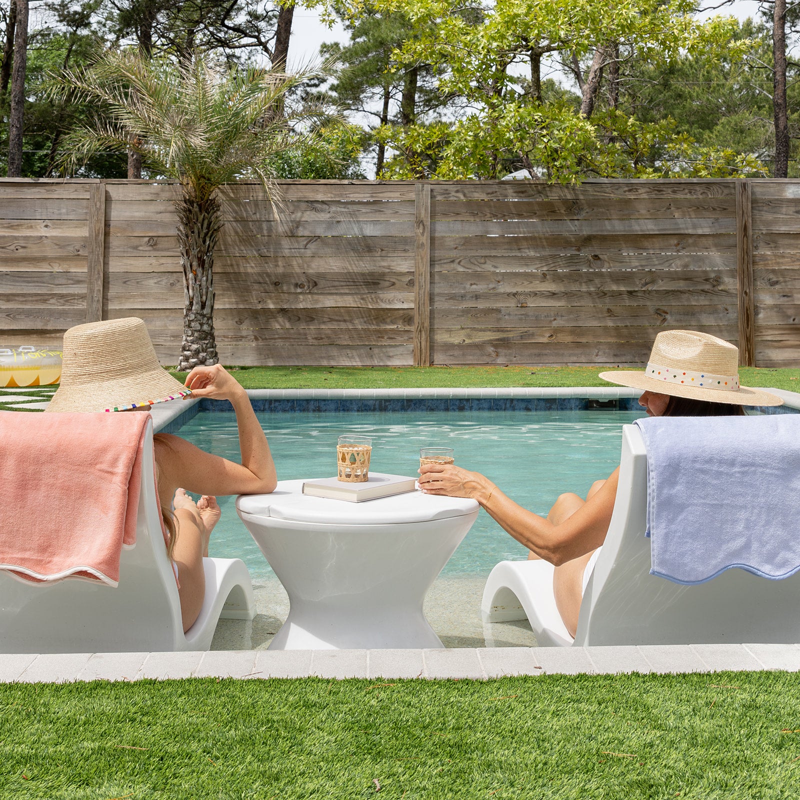 Two women sitting by a pool with drinks, wearing natural woven hats and coral and blue hydrangea colored towels.